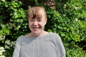 Image of our supported person, Tracy, smiling to the camera with greenery behind her.